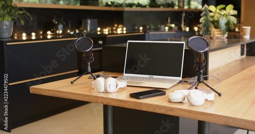 Podcast setup is occupying wooden communal table with open laptop, mics, headphones, phone