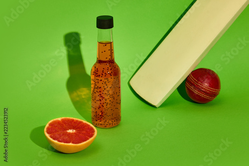 Fotografie Bottled seed drink beside cricket bat, red ball and grapefruit on green background