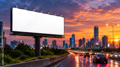 Large blank billboard template standing on a highway with a busy city skyline during a beautiful golden sunset