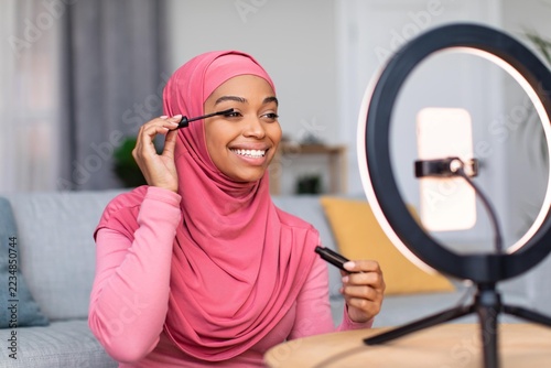 Obraz na plátně Smiling Young African American Woman Applying Mascara With Brush In Front Of Ring Light, Sitting At Home