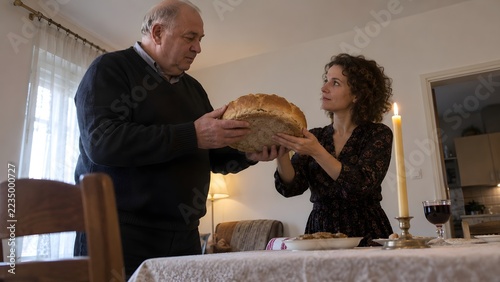 An older man and a woman stand at a table, jointly holding a freshly baked loaf of bread in a domestic setting