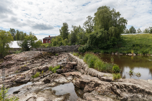 Old stone bridge crosses a rocky riverbed with a red wooden barn on the bank.