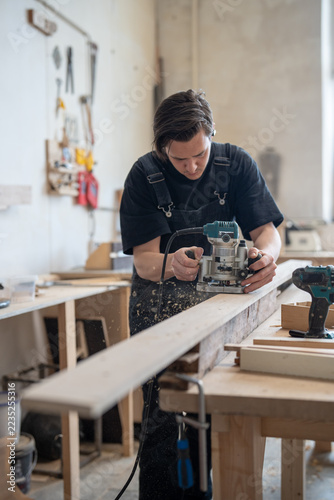 Obraz na plátně Young craftsman guides handheld router along wooden board as sawdust scatters from cutting area, machining timber edge during furniture making, joinery production and professional workshop work