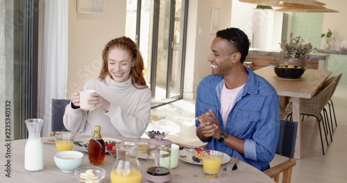 Diverse couple sitting at dining table and laughing over breakfast with white mug, juice glasses