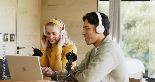 Couple sitting at table in cabin, wearing white headphones and recording with laptop plus mics
