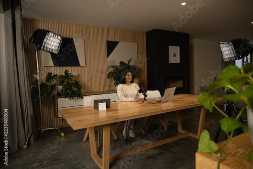 Indian woman wearing sweater and jeans, sitting in home studio hosting podcast with laptop mic