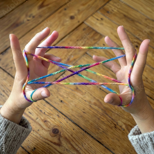 Two hands stretch a multicolored string into a cat's cradle pattern against a warm, rustic wooden floor backdrop