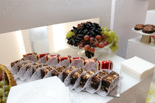 Chocolate pastries with nuts and sponge cake rolls with berry filling arranged beside a fresh grape display.