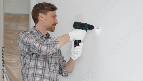 Obraz na plátně Middle aged man worker wearing grey checked shirt and white gloves, is drilling hole in a wall with cordless drill during home renovation work