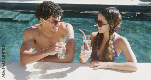 Happy diverse couple in sunglasses standing in sunny swimming pool with drinks