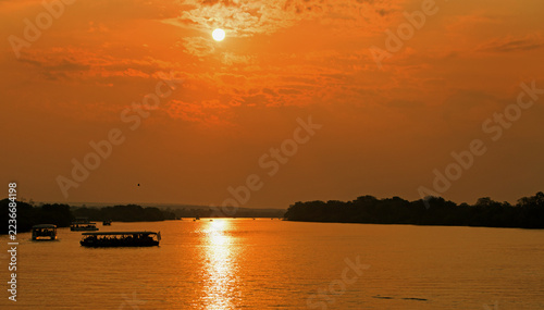 Sunset over the Zambezi River, with bright orange sky with sun reflecting in the water, with passenger tour boats. Zimbabwe, Africa