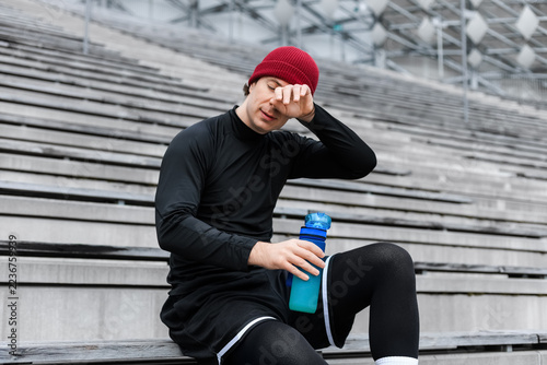 Obraz na plátně Tired runner wiping sweat after cardio workout, man seated on stadium steps with