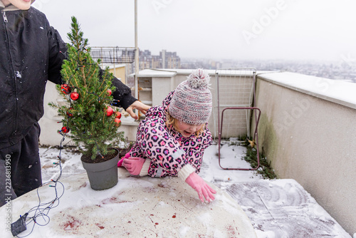 Child draws in snow beside a decorated tree on a rooftop