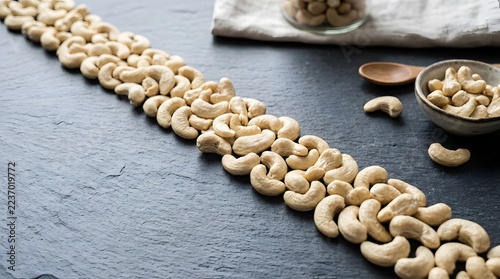 Raw cashew nuts arranged in a flowing wave on a dark slate background with wooden spoon and bowl