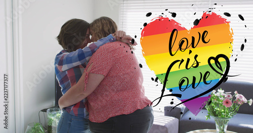 Image of rainbow heart and love is love over lesbian couple embracing at home
