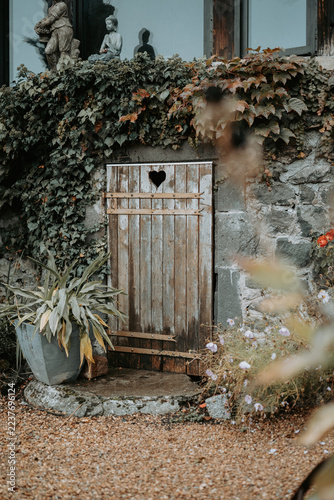 Petite porte en bois avec ouverture en forme de cœur dans un mur en pierre recouvert de lierre