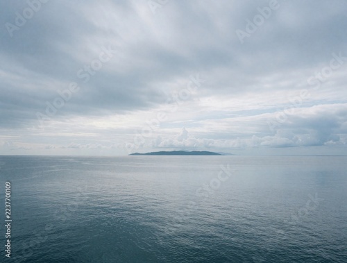 Calm sea horizon under overcast cloudy sky. Distant island sits on quiet ocean surface. Tranquil seascape with misty blue water, grey atmosphere. Minimal nature landscape provides peaceful background