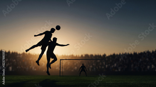 Silhouette of soccer players jumping to header a football in front of a goalpost during sunset in a packed stadium